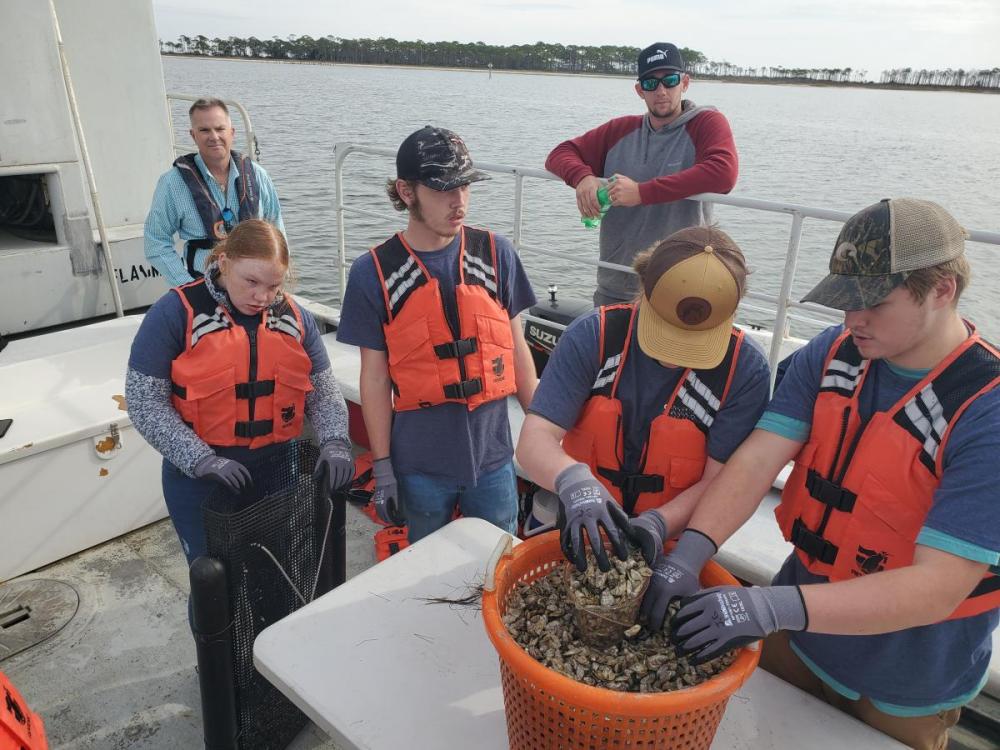 Students and adults on boat wearing life vests. Two students have hands in a bucket of oysters.