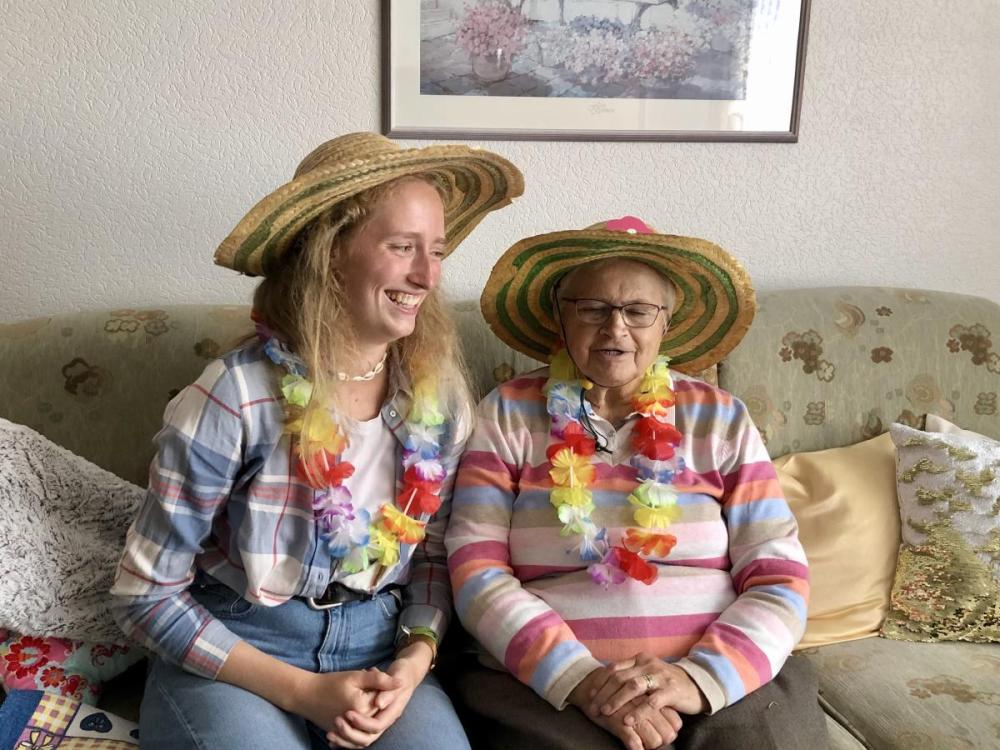 Carolin Ellerkamp and her grandmother Marianne Borgers sit on a couch together, both smiling and wearing straw hats and flower leis.