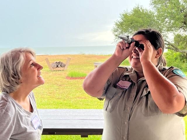 Woman on the left looks up at the woman on the right who is holding up a monoscope-shaped device. In the background is the ocean, sky, and a grassy field.