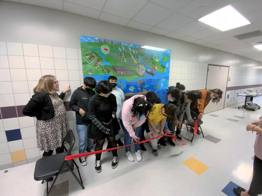 Two teachers and a group of students cut red ribbon in a ceremonial unveiling of mural
