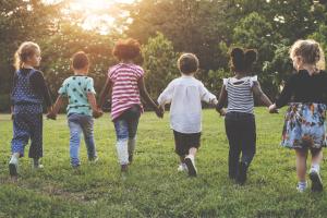 Six children hold hands in a line walking towards trees in a field as the sun peeks through the canopy.