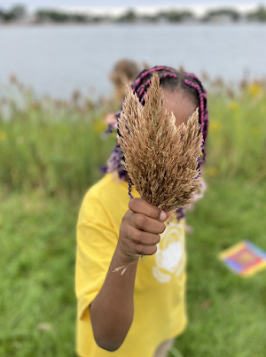Photo of a young Black child, their face hidden behind a bushel of yellowed reeds that are grasped in the child’s hand.