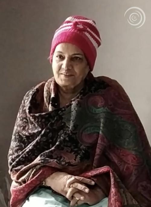 A woman in a flowered warp and pink and white knit hat sits with hands folded. 